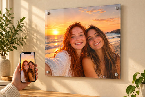 Phone held in front of a wall-mounted acrylic print of the same beach sunset selfie, showing a phone photo turned into wall art